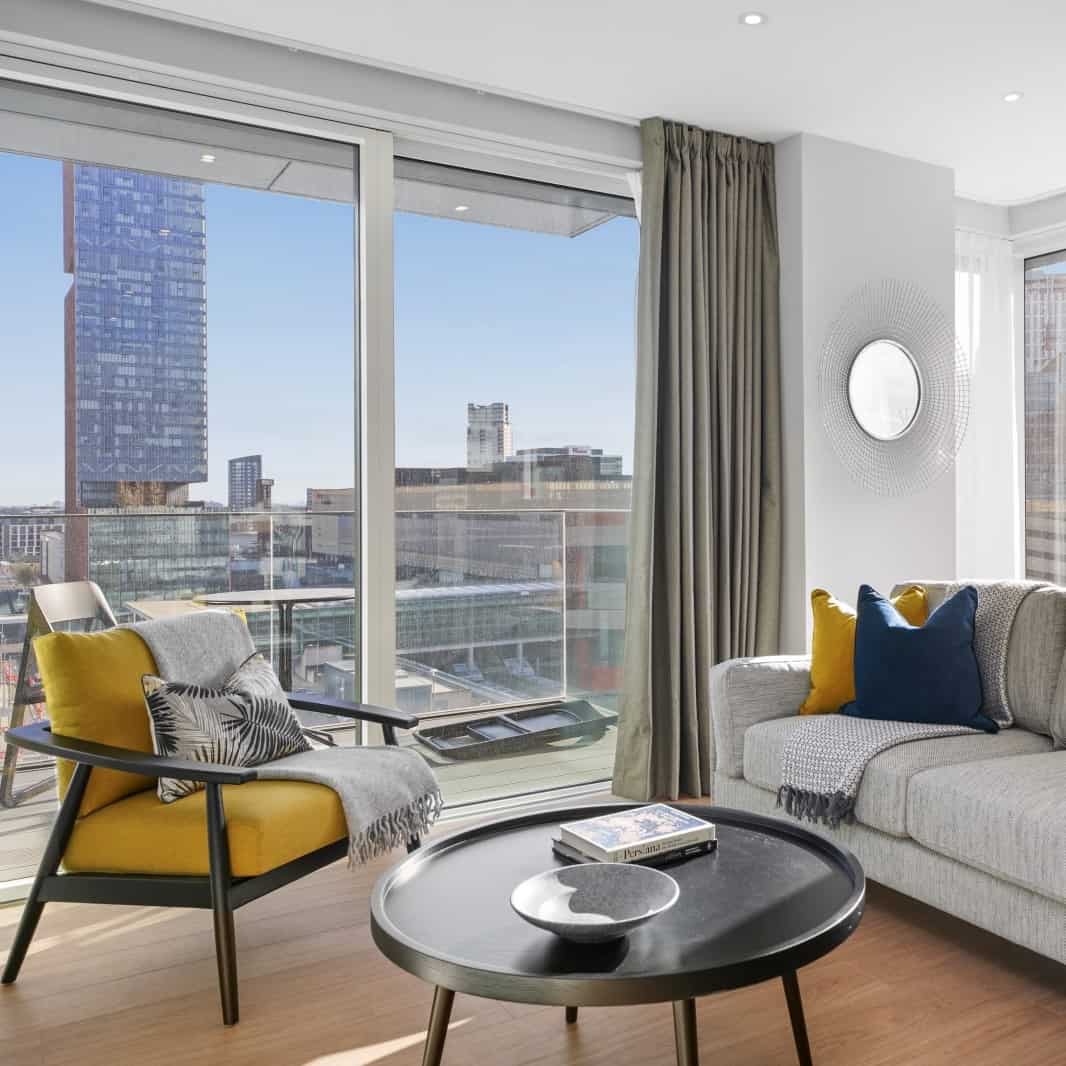 A Skylark apartment living area with a long sofa and armchair, floor to ceiling window views of The Stratford hotel and restaurant