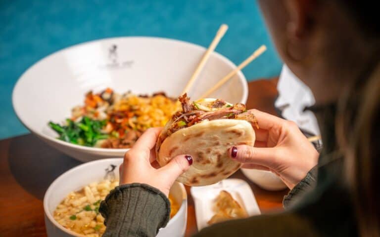 Hands holding a traditional Xi'an Murger with other food in the background at Murger Han restaurant