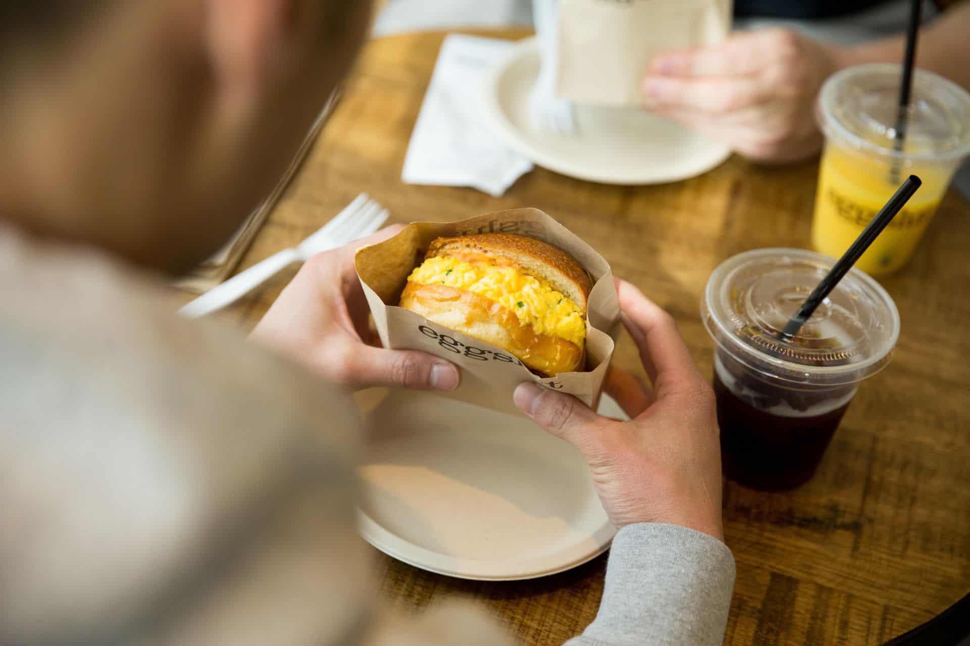 Hands holding an egg sandwich at an Eggslut restaurant with drinks on the table