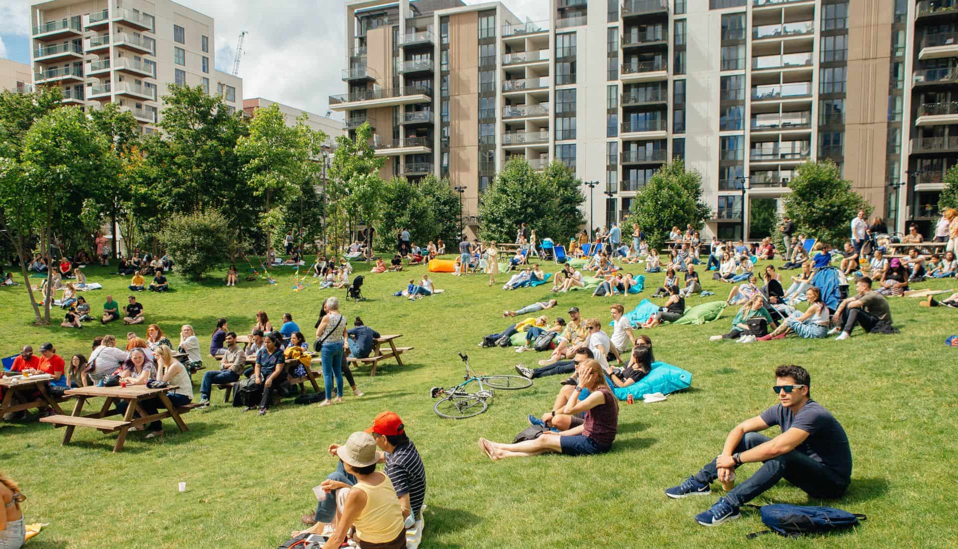 People on the grass of Victory Park at a summer community event with East Village buildings in the background