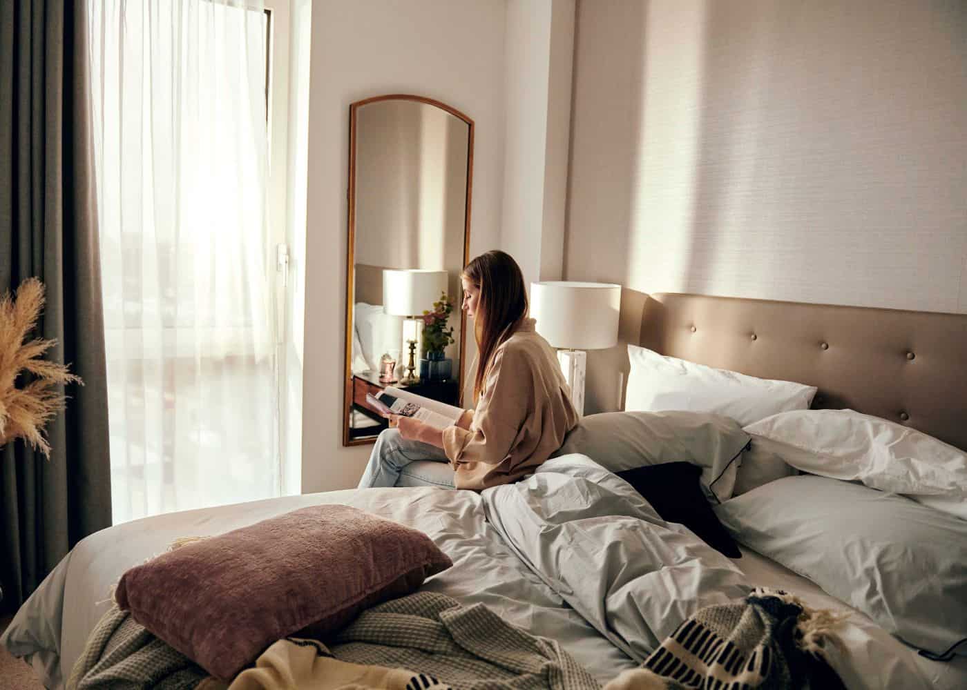 Woman sat on her unmade bed reading a magazine with the morning light shining into the apartment bedroom