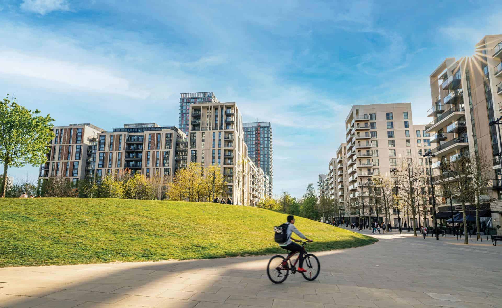 A cyclist passing through East Village by Victory Park on a sunny day with apartment buildings in the background